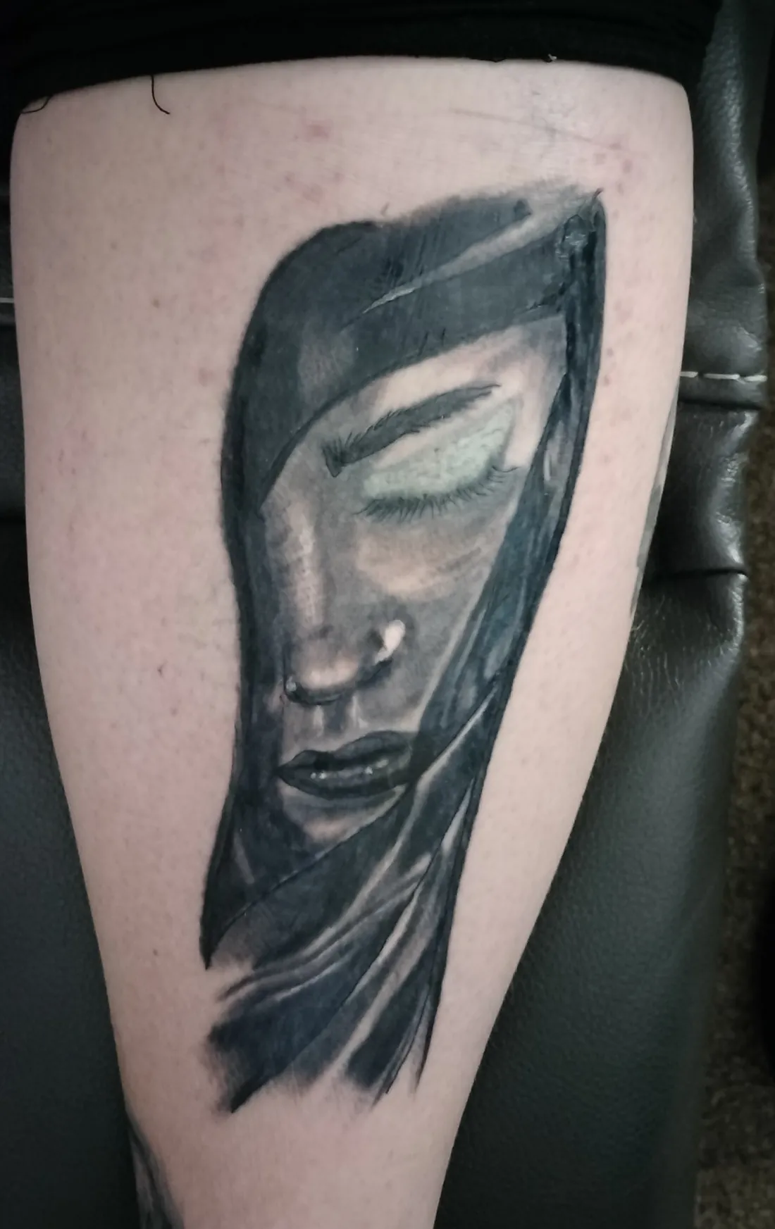 A woman with her eyes closed under a dark hood, black and grey, tattooed on a calf
